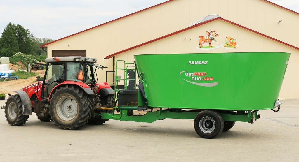 Wóz paszowy DUO firmy Samasz przeznaczony dla dużych gospodarstw. Widok boczny maszyny z ciągnikiem na tle obory