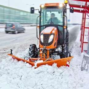 Pomarańczowy traktor rolniczy wraz z pomarańczowym spychem do śniegu CITY 180 firmy Samasz podczas odśnieżania zatoczkę przy przystanku www.korbanek.pl